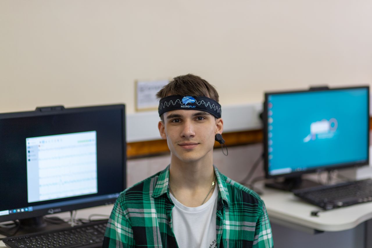 Portrait of a young man in a plaid shirt wearing a Neuroplay headband with computers in the background.